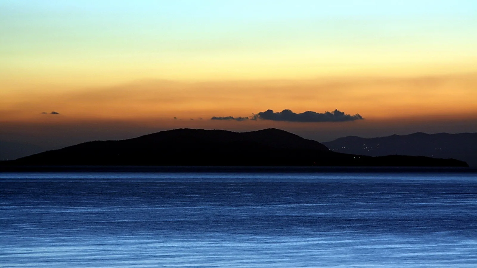 Dunkle Silhouette einer Insel vor ruhigem blauem Meer, darüber ein orangefarbener Abendhimmel mit einem schmalen Wolkenband und Bergen im Hintergrund.