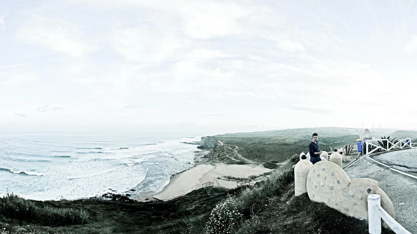 Panorama einer Steilküste mit Strand und Brandung, links das offene Meer, rechts ein Mann auf einer Betonskulptur an einem weißen Geländer.