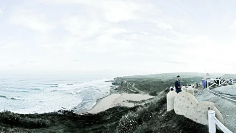 Panorama einer Steilküste mit Strand und Brandung, links das offene Meer, rechts ein Mann auf einer Betonskulptur an einem weißen Geländer.