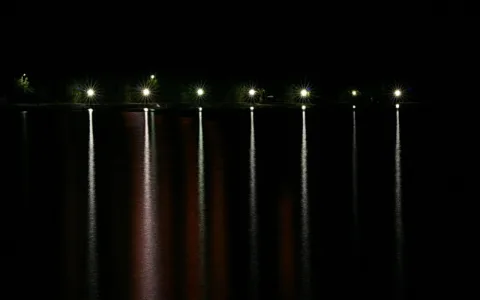 Eine Reihe beleuchteter Laternen über einem dunklen Wasser, deren Lichter sich als helle vertikale Streifen in der Wasseroberfläche spiegeln, vor schwarzem Nachthimmel.