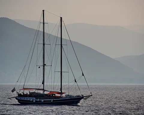 Ein zweimastiges Segelboot mit sichtbarer Takelage fährt auf ruhigem Meer, im Hintergrund liegen blau-graue Bergketten im Dunst.