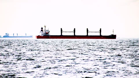 Rotes Frachtschiff mit weißem Aufbau und mehreren schwarzen Kränen fährt über bewegtes Meer, im Hintergrund ein zweites Schiff im Dunst.