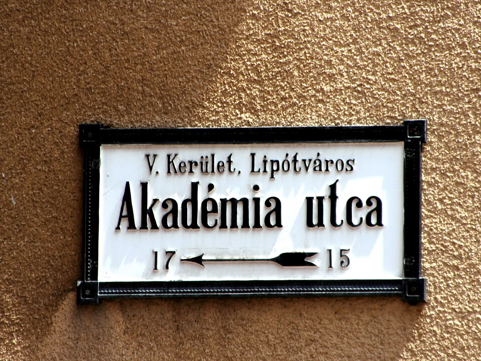 Weißes Straßenschild mit schwarzem Rahmen auf ockerfarbener Putzwand, Aufschrift V. Kerület, Lipótváros und Akadémia utca, darunter Hausnummern 17 und 15 mit einem Pfeil
