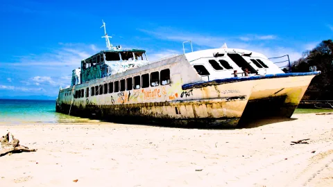 Ein verrostetes, bemaltes Fährschiff mit zwei Rümpfen liegt auf weißem Sandstrand, im Hintergrund türkisfarbenes Meer und blauer Himmel.