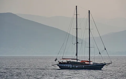 Eine zweimastige Segelyacht fährt auf ruhigem Meer vor blaugrauen Bergen, am Heck weht eine griechische Flagge.