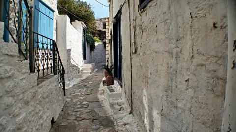 Schmale Gasse mit Kopfsteinpflaster zwischen weiß getünchten Steinmauern, links ein schmiedeeisernes Geländer vor blauen Fensterläden, rechts eine dunkle Tür und ein Blumentopf am Treppenabsatz.
