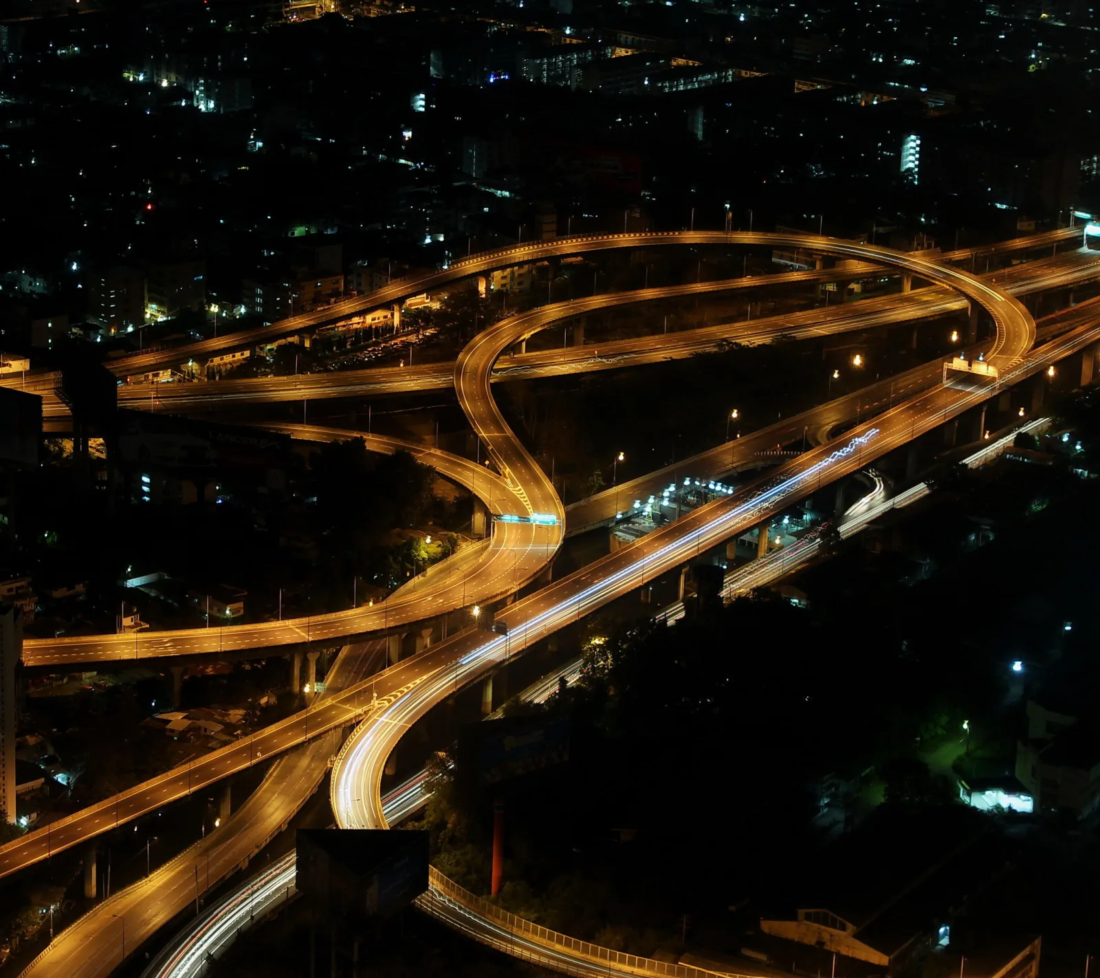 Mehrstöckiges Autobahnkreuz bei Nacht mit geschwungenen Fahrbahnen, auf denen Lichtspuren von Fahrzeugen zu sehen sind, im Hintergrund eine dunkle Stadt mit einzelnen Lichtern