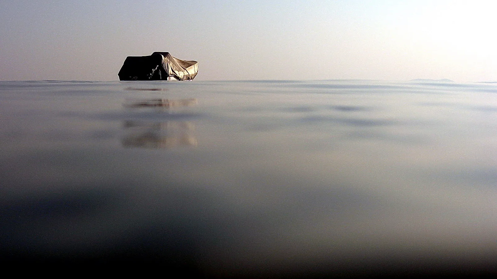 Ein mit einer Plane abgedecktes Boot liegt auf ruhigem Wasser, darüber ein heller, diesiger Himmel.