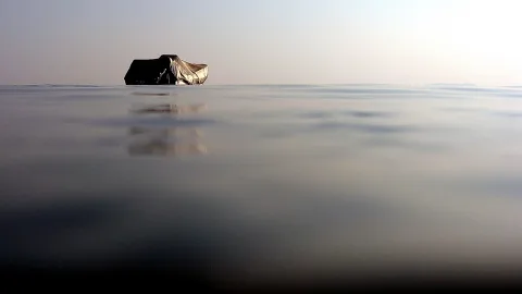 Ein mit einer Plane abgedecktes Boot liegt auf ruhigem Wasser, darüber ein heller, diesiger Himmel.