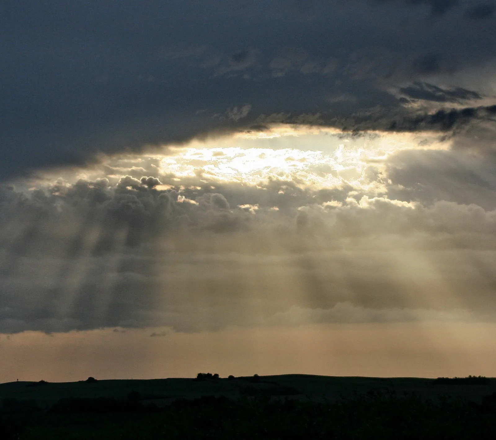 Sonnenlicht bricht als Strahlenbündel durch eine Lücke in dunklen Wolken und beleuchtet die Wolkendecke über einer dunklen, flachen Hügellandschaft am Horizont.