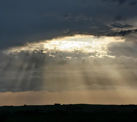 Sonnenlicht bricht als Strahlenbündel durch eine Lücke in dunklen Wolken und beleuchtet die Wolkendecke über einer dunklen, flachen Hügellandschaft am Horizont.