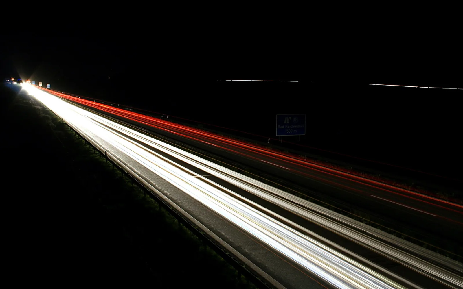 Nächtliche Autobahn mit langen Lichtspuren: weiße Scheinwerfer auf der linken Fahrbahn, rote Rücklichter auf der rechten, dahinter ein blaues Hinweisschild vor schwarzem Himmel.