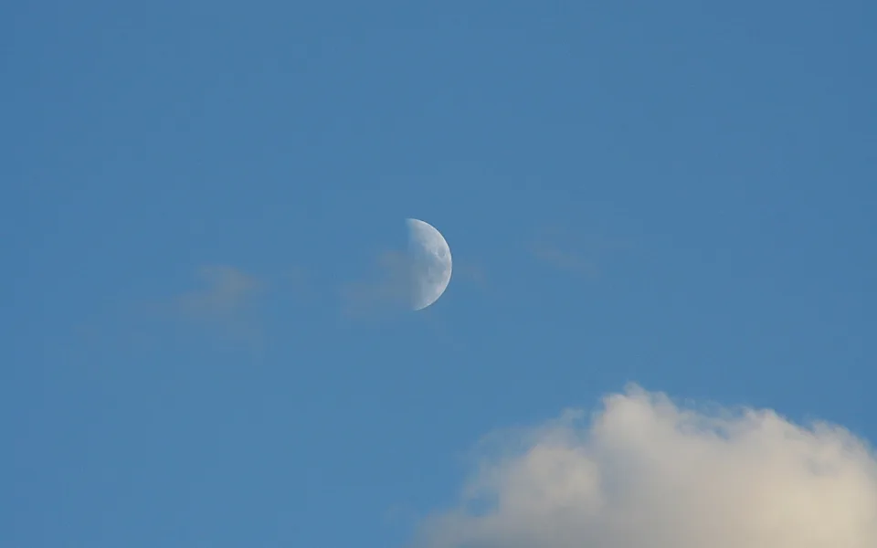 Halbmond am hellblauen Himmel, rechts unten eine weiße Haufenwolke, sonst klarer Himmel.