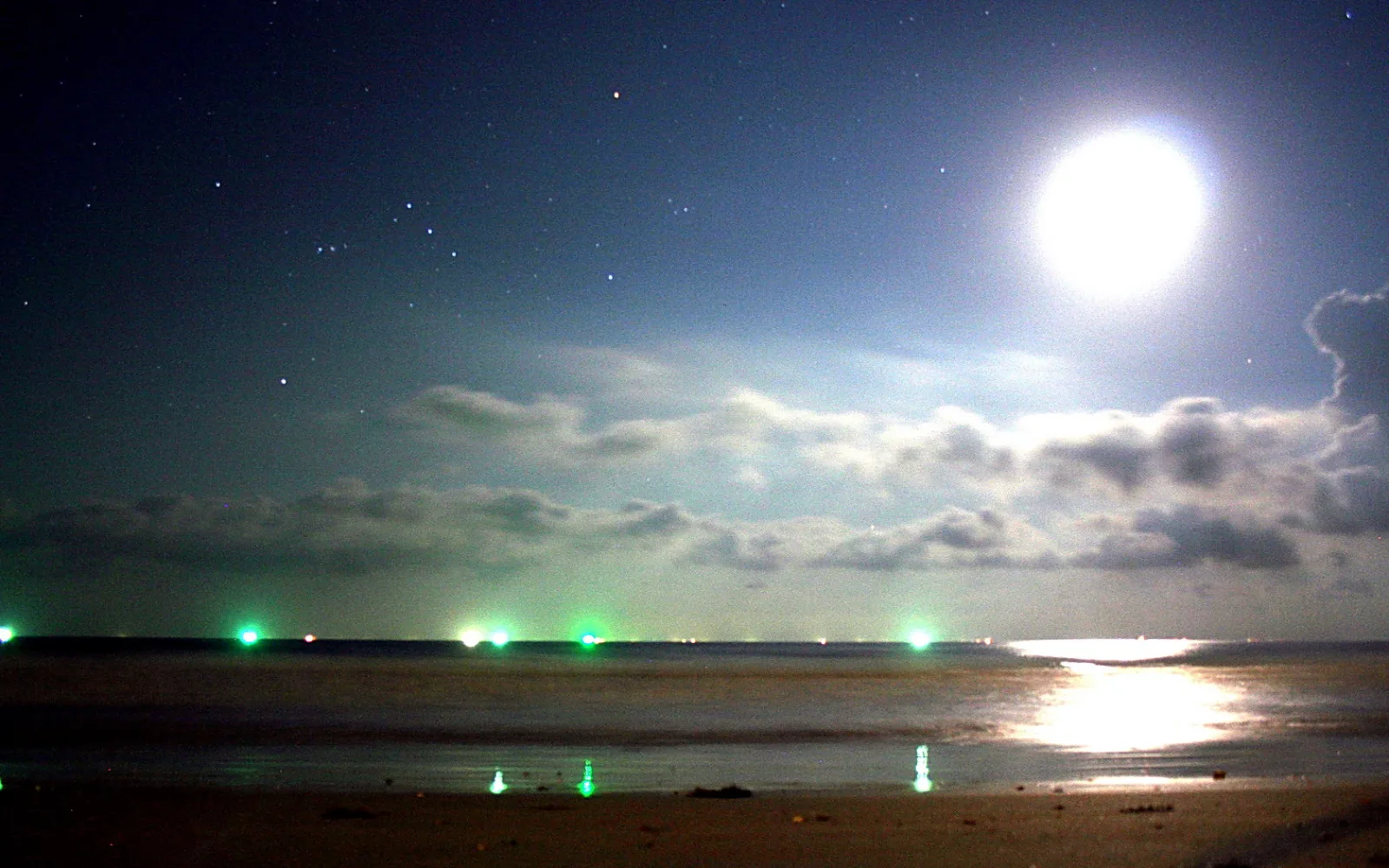 Nächtlicher Strand unter einem hellen Vollmond mit Schleierwolken und Sternen am Himmel, im Wasser spiegelt sich das Mondlicht, am Horizont blinken grüne Lichter.