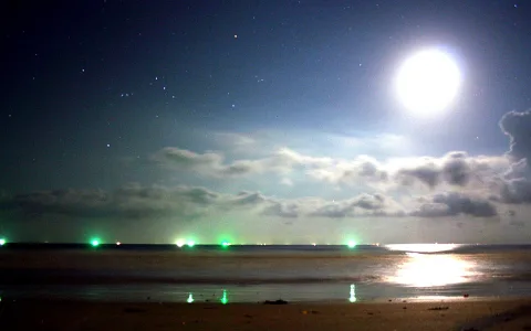 Nächtlicher Strand unter einem hellen Vollmond mit Schleierwolken und Sternen am Himmel, im Wasser spiegelt sich das Mondlicht, am Horizont blinken grüne Lichter.