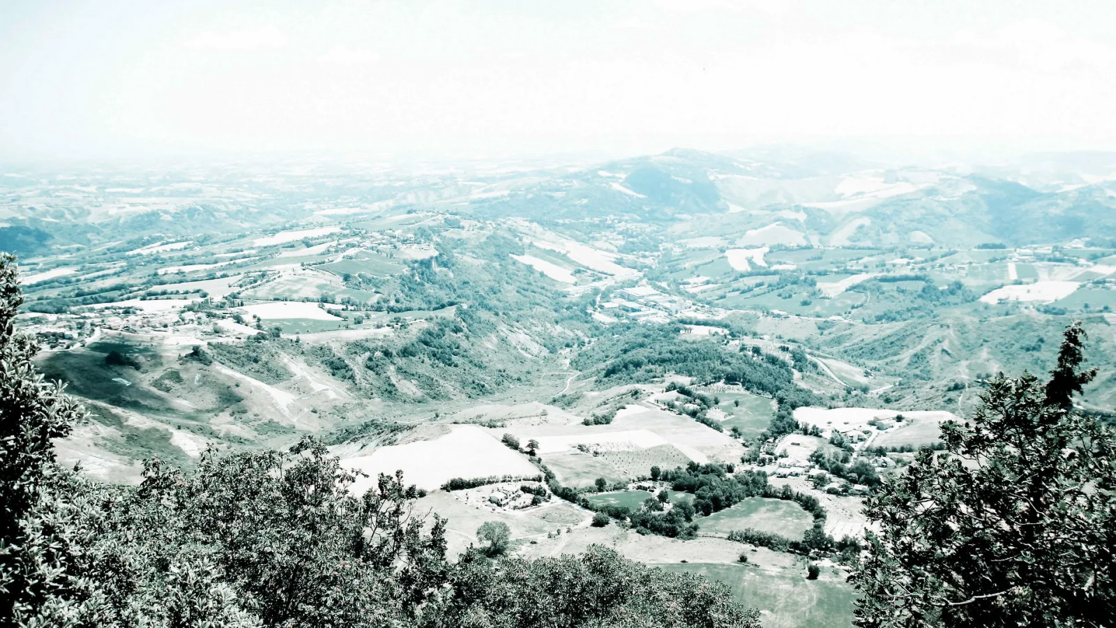 Blick von einer Anhöhe über weite, hügelige Landschaft mit Feldern, kleinen Ortschaften und Baumgruppen im Vordergrund, überzogen von hellem Dunst.