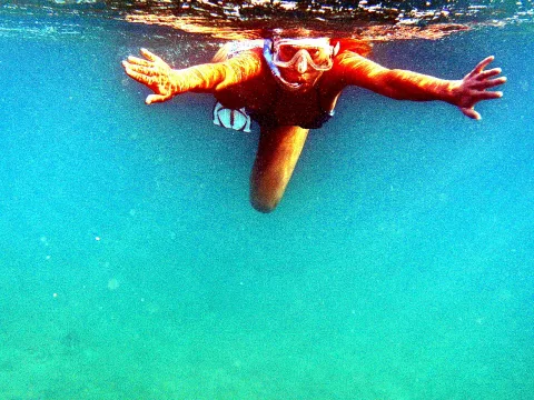 Schnorchelnde Person mit Taucherbrille unter Wasser, beide Arme weit ausgebreitet, im türkisfarbenen Meer, Wasseroberfläche oben im Bild.