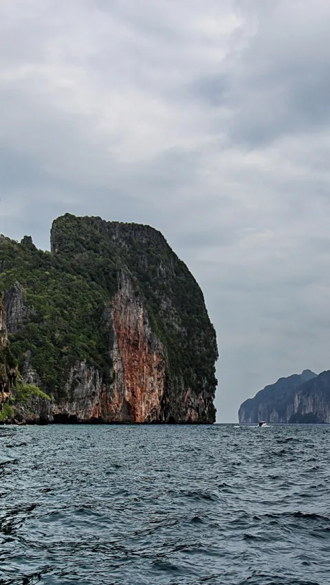 Steil aufragende, bewachsene Kalkfelsen über dem Meer, rechts im Hintergrund ein weiterer Felsrücken und ein kleines Boot unter bewölktem Himmel.