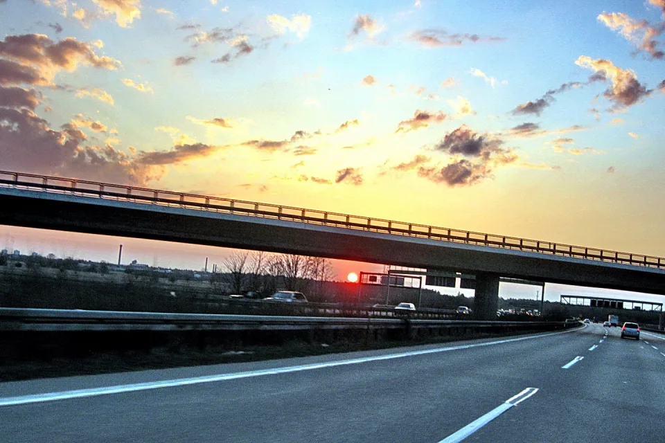 Eine Autobahn führt unter einer Brücke hindurch, im Hintergrund geht die Sonne über einer Landschaft mit Wolken unter, auf der Fahrbahn fahren Autos.