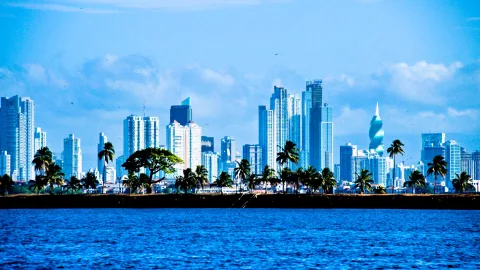 Hochhäuser der Skyline von Panama-Stadt hinter einer dunklen Uferlinie mit Palmen, davor blaues Meer unter leicht bewölktem Himmel.