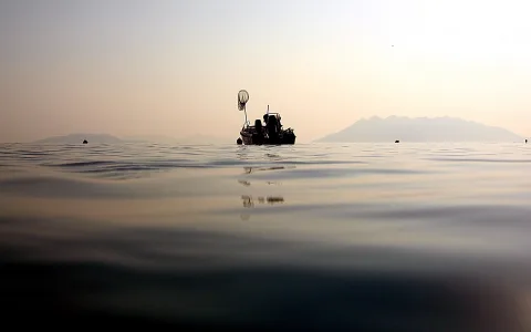 Ein kleines Fischerboot mit einem Netz am Mast schwimmt im Morgenlicht auf ruhigem Wasser, im Hintergrund liegen flache Berge im Dunst.