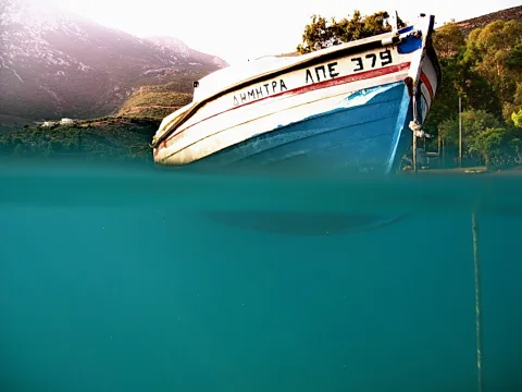 Weiß-blaues Fischerboot mit griechischer Aufschrift, zur Hälfte über der Wasseroberfläche, davor bewaldete Berge