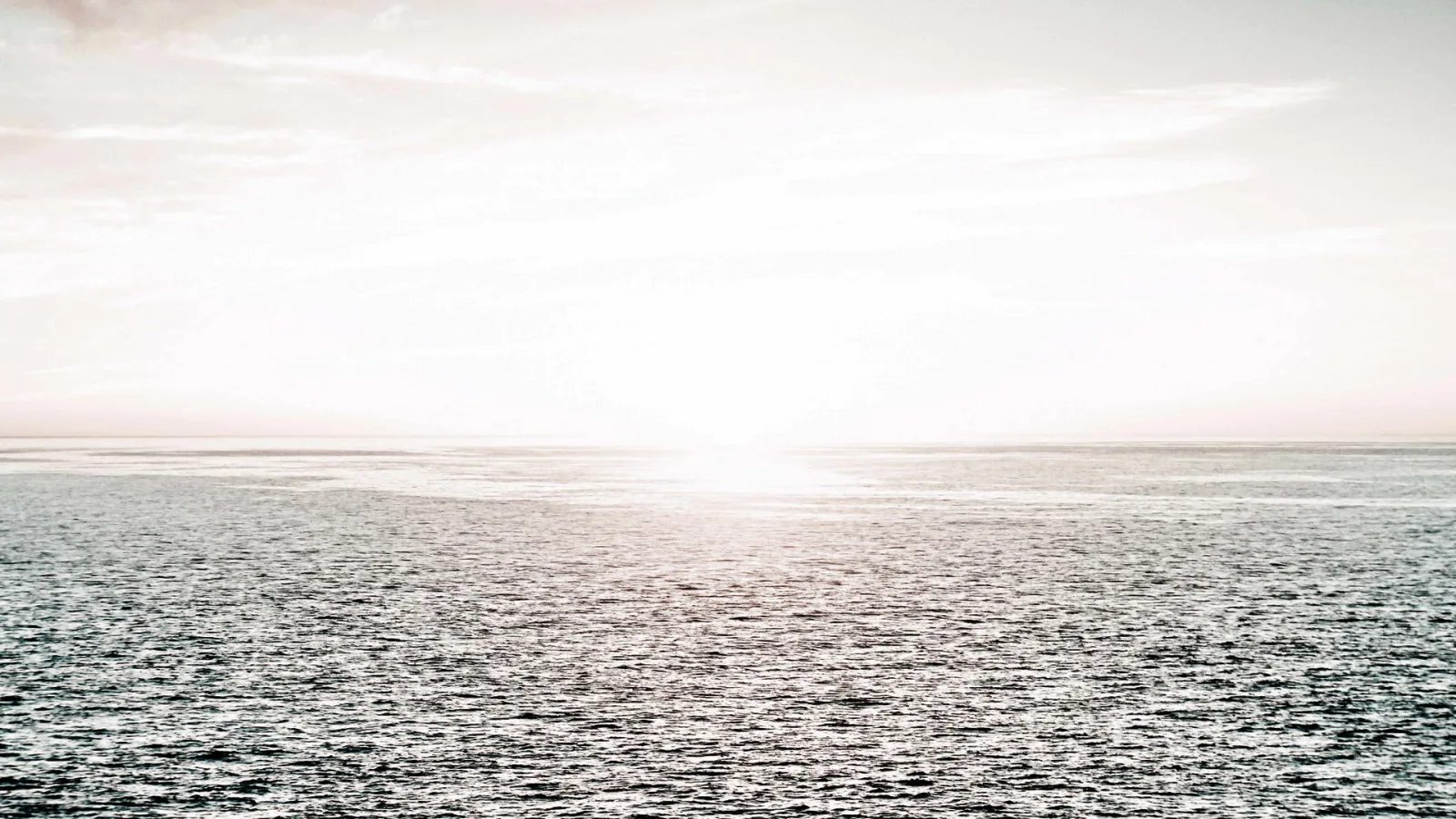 Ruhiges Meer unter einem hellen, dunstigen Himmel. Die tief stehende Sonne spiegelt sich als gleißender Lichtfleck auf der Wasseroberfläche am Horizont.