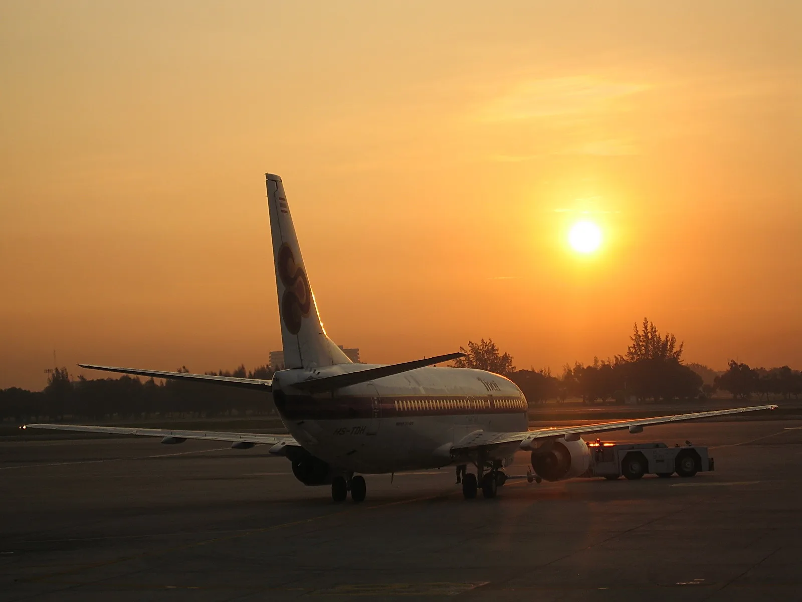 Ein Passagierflugzeug von hinten auf dem Vorfeld, im Gegenlicht der tief stehenden Sonne, daneben ein Schlepper.