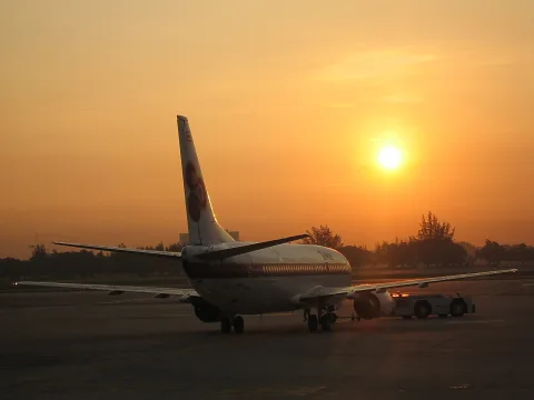 Ein Passagierflugzeug von hinten auf dem Vorfeld, im Gegenlicht der tief stehenden Sonne, daneben ein Schlepper.