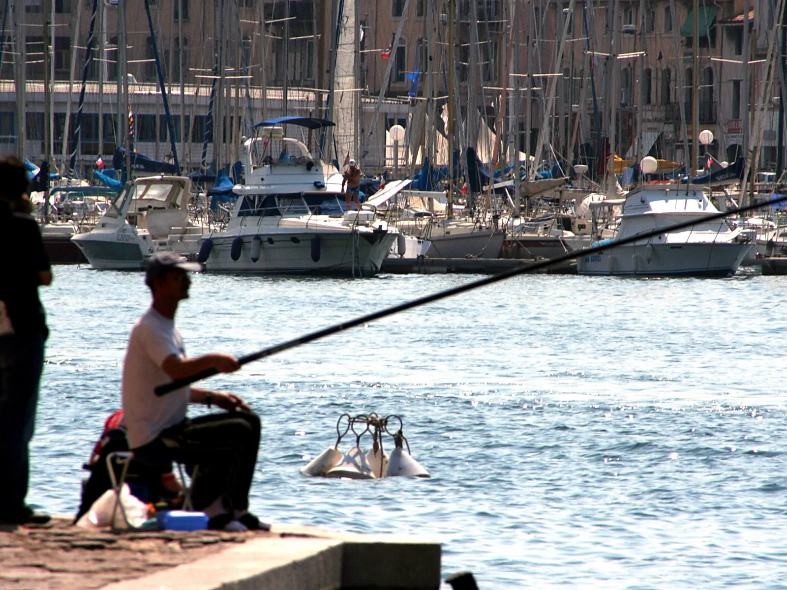 Ein Angler sitzt auf einer Kaimauer und hält eine lange Angelrute über das Wasser. Im Hintergrund liegen viele Segelboote und Motoryachten dicht nebeneinander im Hafen, dahinter stehen Gebäude.