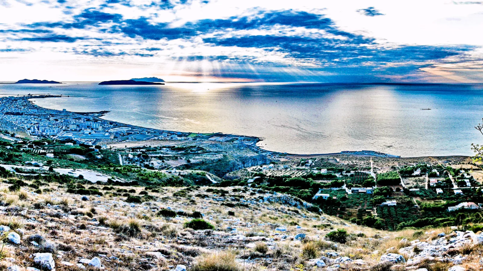 Weite Aussicht von einem trockenen Hügel auf eine Küstenstadt, das ruhige Meer mit Inseln am Horizont und Sonnenstrahlen durch die Wolken.