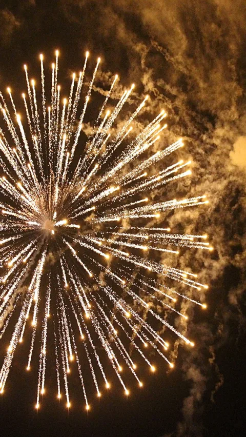 Ein goldweiß leuchtendes Feuerwerk mit strahlenförmig auseinanderlaufenden Funken vor dunklem Nachthimmel, rechts ziehen Rauchschwaden vorbei.