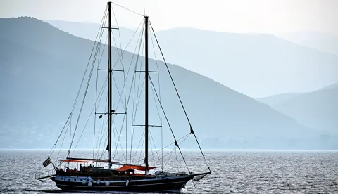 Zweimastiges Segelboot mit griechischer Flagge auf ruhigem Meer vor blau getönten Bergen.