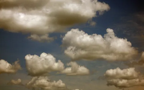 Mehrere weiße Kumuluswolken mit grauen Schattenpartien vor einem dunkelblauen Himmel, unten rechts ein warmer Gelbton.