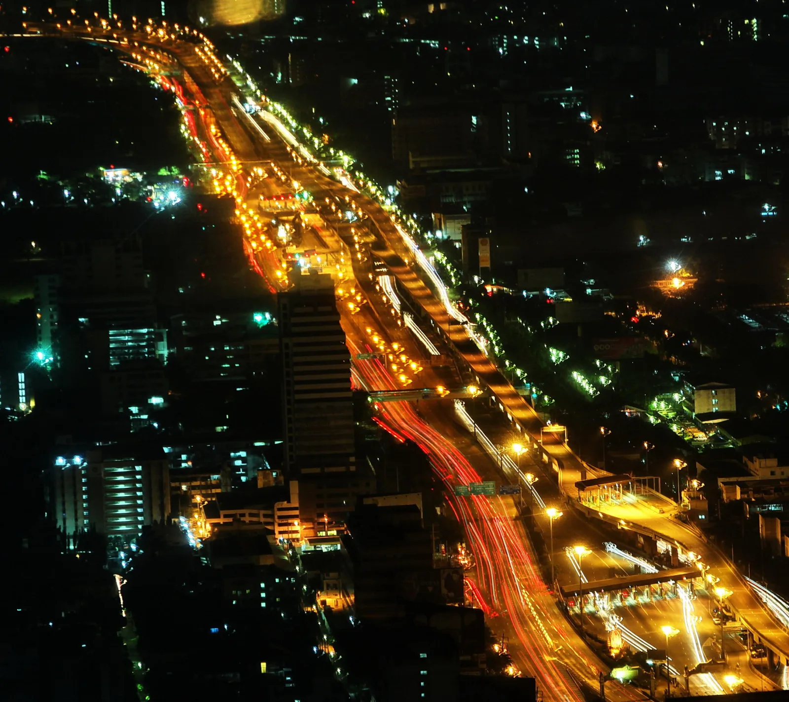 Luftaufnahme einer mehrspurigen Stadtautobahn bei Nacht mit gelben Straßenlaternen und roten und weißen Lichtspuren von Fahrzeugen, umgeben von dicht bebauter Stadt mit beleuchteten Hochhäusern.
