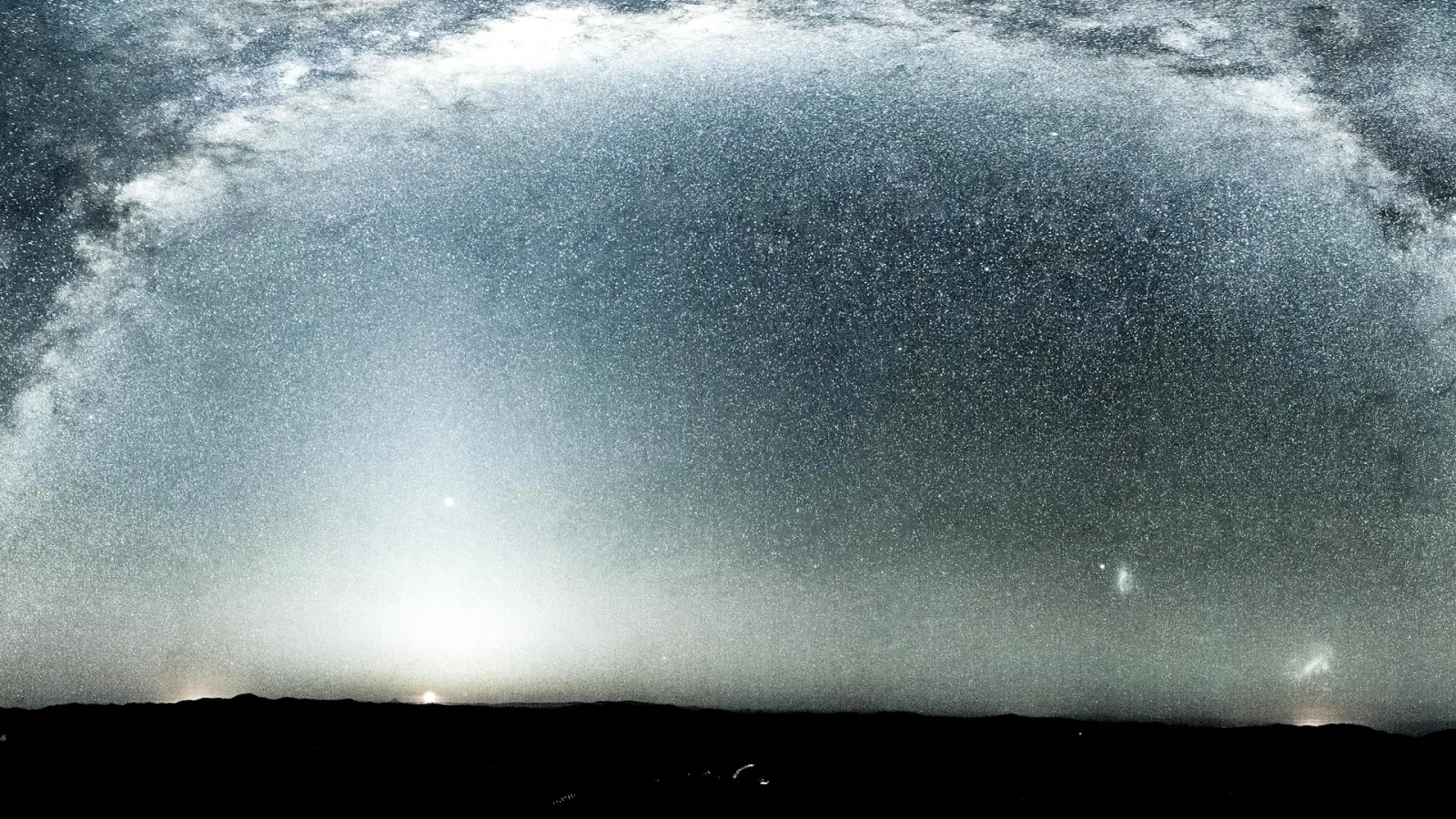Sternenreicher Nachthimmel mit einem hellen Bogen der Milchstraße über einer dunklen Hügelkette, am Horizont leuchtet ein diffuser Lichtschein.