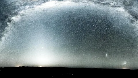 Sternenreicher Nachthimmel mit einem hellen Bogen der Milchstraße über einer dunklen Hügelkette, am Horizont leuchtet ein diffuser Lichtschein.