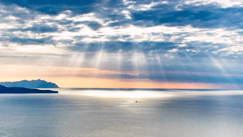 Sonnenstrahlen brechen durch aufgerissene Wolken und fallen auf eine glatte Meeresfläche, links ragen dunkle Küstenhügel ins Wasser, in der Mitte ist ein winziges Boot zu sehen.