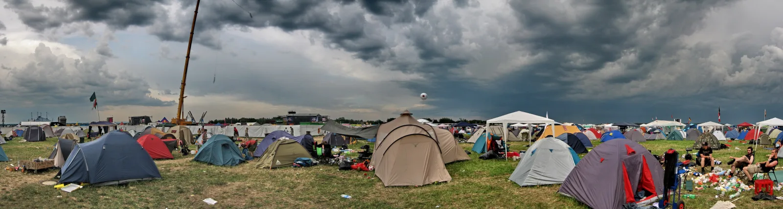 Panoramablick auf einen Festivalzeltplatz auf grüner Wiese unter dramatischen dunklen Gewitterwolken. Zahlreiche Zelte in Blau, Beige, Grau und Rot stehen verstreut, im Hintergrund ragt ein Kran auf, und rechts sitzen einige Menschen auf Campingstühlen.