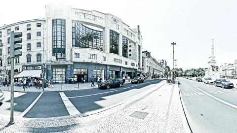 Breites Panorama einer Straßenkreuzung mit Pflaster, Altbauten mit großen Fenstern, Autos und einer Säule rechts im Bild, in Schwarzweiß mit leichtem Blaustich.
