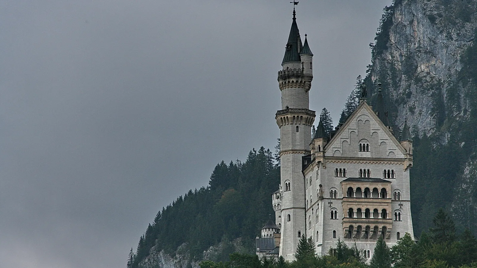 Schloss Neuschwanstein mit hellen Türmen vor einer steilen, bewaldeten Felswand unter grauem Himmel.