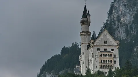Schloss Neuschwanstein mit hellen Türmen vor einer steilen, bewaldeten Felswand unter grauem Himmel.