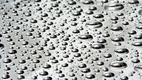 Viele kleine und größere Wassertropfen liegen dicht gedrängt auf einer grau glänzenden Oberfläche, die Lichter spiegeln sich in den Tropfen.