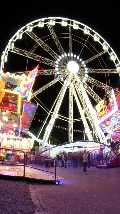 Ein beleuchtetes Riesenrad vor schwarzem Nachthimmel, links und rechts bunt leuchtende Fahrgeschäfte und Buden, davor Menschen auf einem Weg.