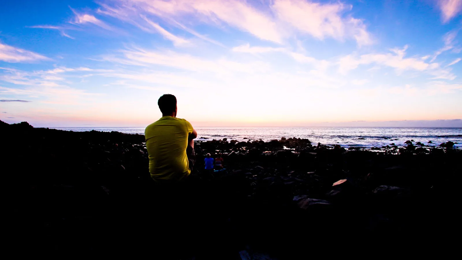 Ein Mann in gelbem T-Shirt sitzt auf dunklen Felsen und schaut auf das Meer. Der Himmel leuchtet in Blau und Rosa über dem Horizont.