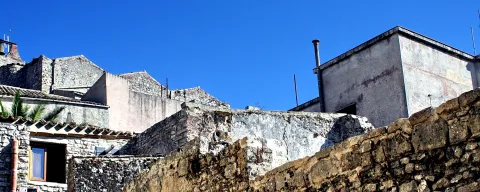 Alte Steinhäuser und verwitterte Mauern unter blauem Himmel, rechts ragt ein Schornstein auf.