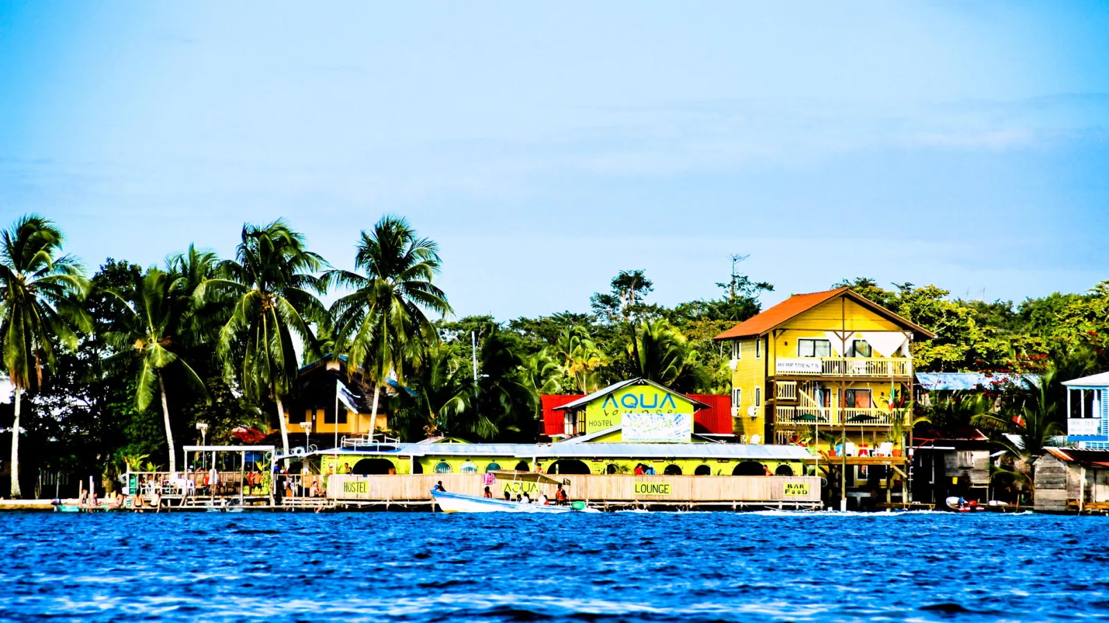 Gelbes Hostel mit Palmen am Ufer von Bocas del Toro, davor blaues Wasser und ein kleines Boot.