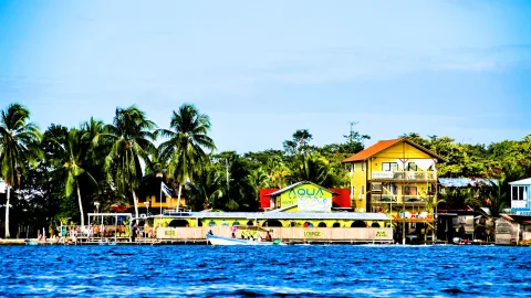 Gelbes Hostel mit Palmen am Ufer von Bocas del Toro, davor blaues Wasser und ein kleines Boot.
