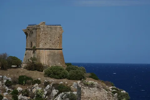 Verwitterter Steinturm auf einer felsigen Küste vor dunkelblauem Meer und klarem Himmel.
