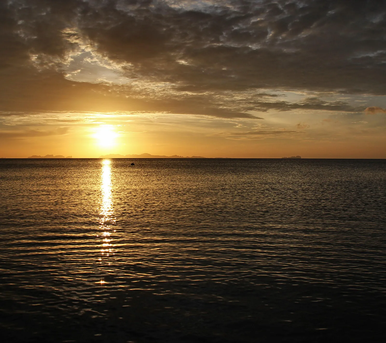 Die tief stehende Sonne spiegelt sich als goldene Bahn auf einer ruhigen Meeresoberfläche, darüber ein dunkles Wolkenband, am Horizont sind Inselsilhouetten zu sehen.
